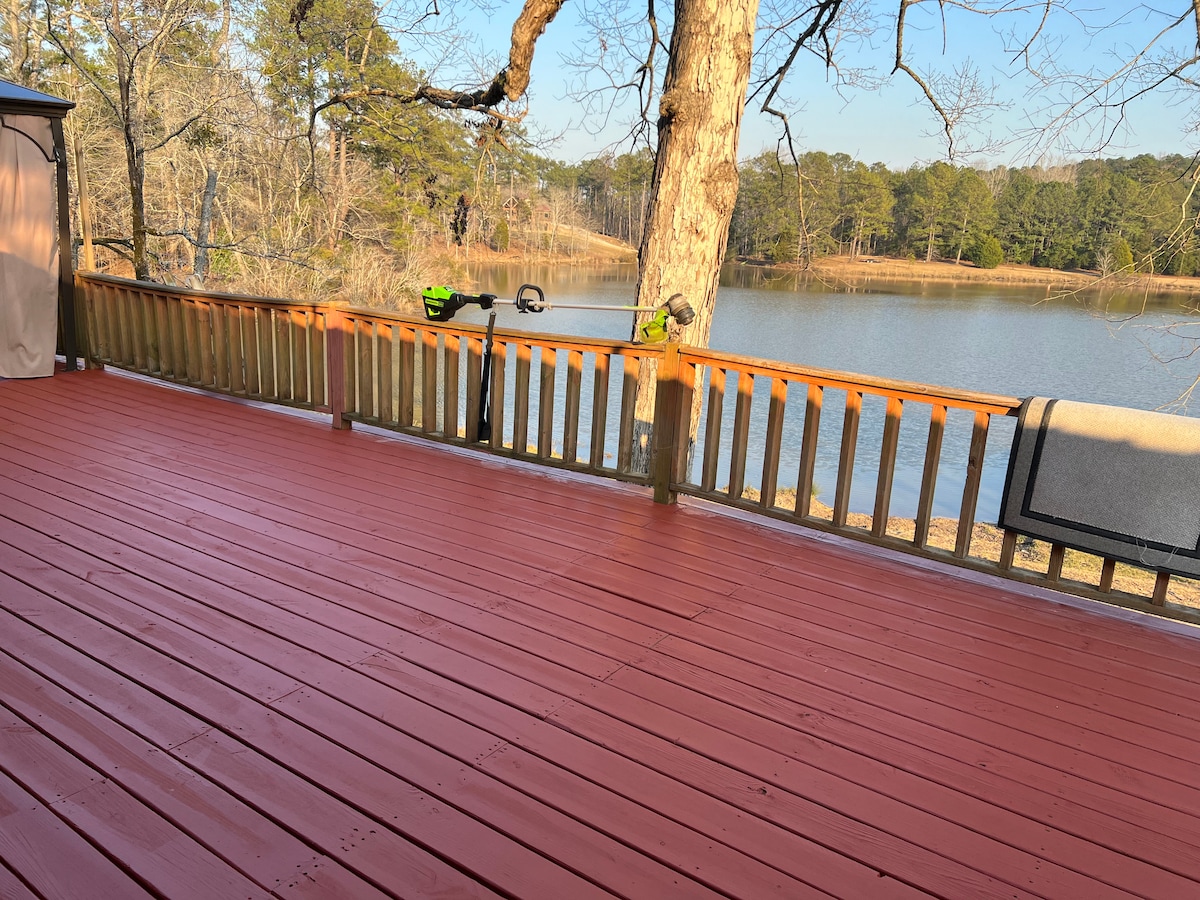 A wooden deck features a vibrant red finish, overlooking a serene body of water surrounded by trees. A grey towel is draped over the railing, while a green tool rests thoughtfully along the edge. The setting suggests a peaceful retreat in nature.