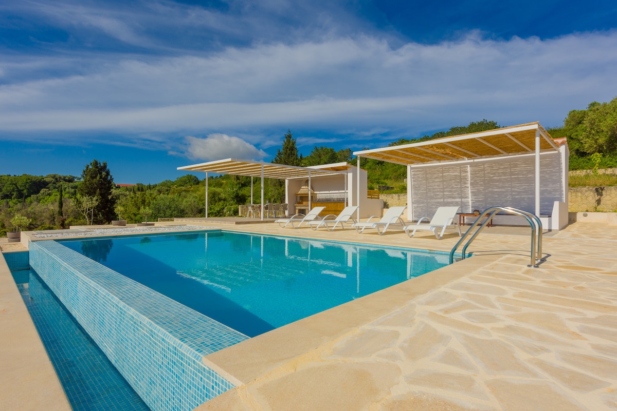 A sparkling outdoor pool is surrounded by a sun deck, featuring several lounge chairs under shaded canopies. The area is framed by lush greenery, with a clear blue sky overhead and soft clouds enhancing the serene environment.