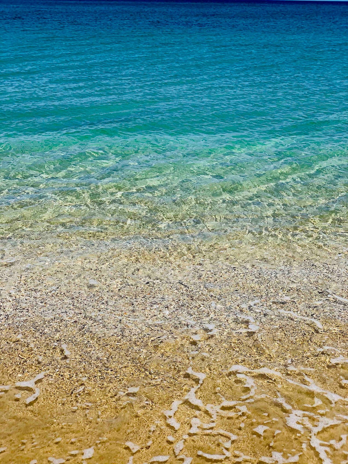 A clear view of calm water gently lapping against a sandy shore is depicted. The water showcases various shades of blue, transitioning from deep hues to lighter tones near the beach, while small waves create a frothy white edge at the shoreline.