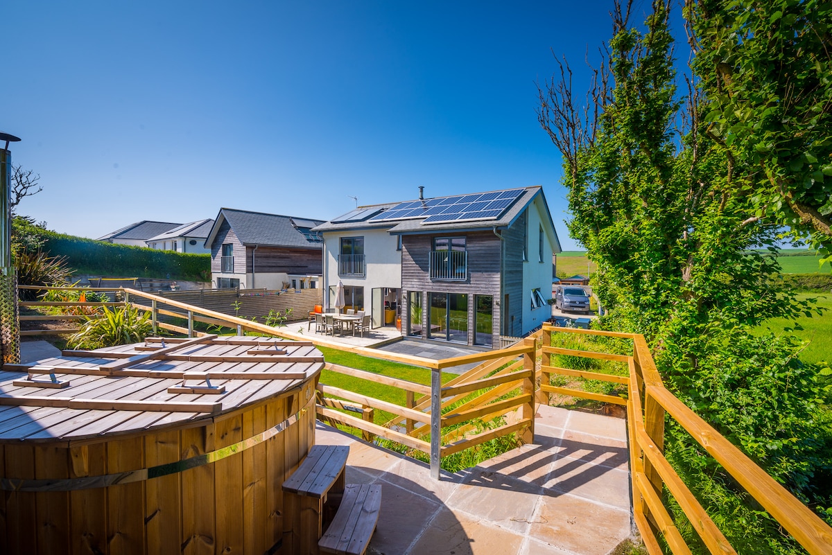 A modern two-story home displays large glass doors that connect the interior to the outdoor patio area. Solar panels are positioned on the roof. A wooden hot tub, surrounded by a railing, is visible in the foreground, set against a backdrop of greenery.