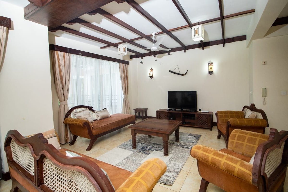 A spacious living room features comfortable seating arrangements with wooden accents. A coffee table is centered on a textured area rug, while a flat-screen TV is positioned on a media unit against a light-colored wall. Natural light enters through sheer curtains.