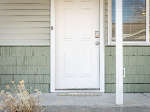 The entryway features a white front door framed by light green siding and a concrete stoop. A welcome mat is positioned at the base of the door, and a small flower arrangement adds a touch of decor. Natural light enters through the nearby window.