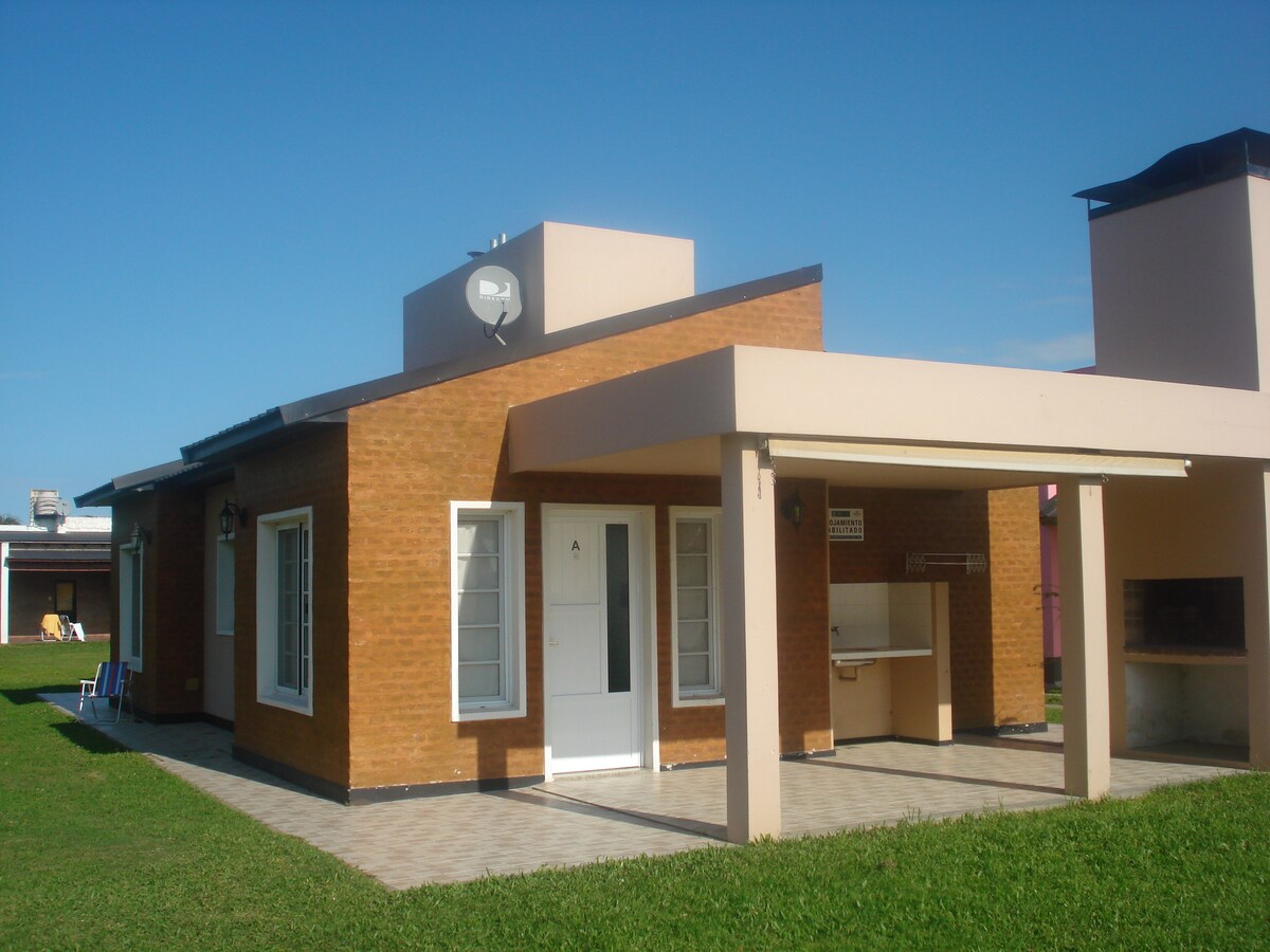 A single-story cabin features a brick façade and a light-colored roof. A covered patio extends from the entrance, providing access to an outdoor grill area. Surrounding green grass adds to the outdoor space, while a clear blue sky enhances the overall setting.