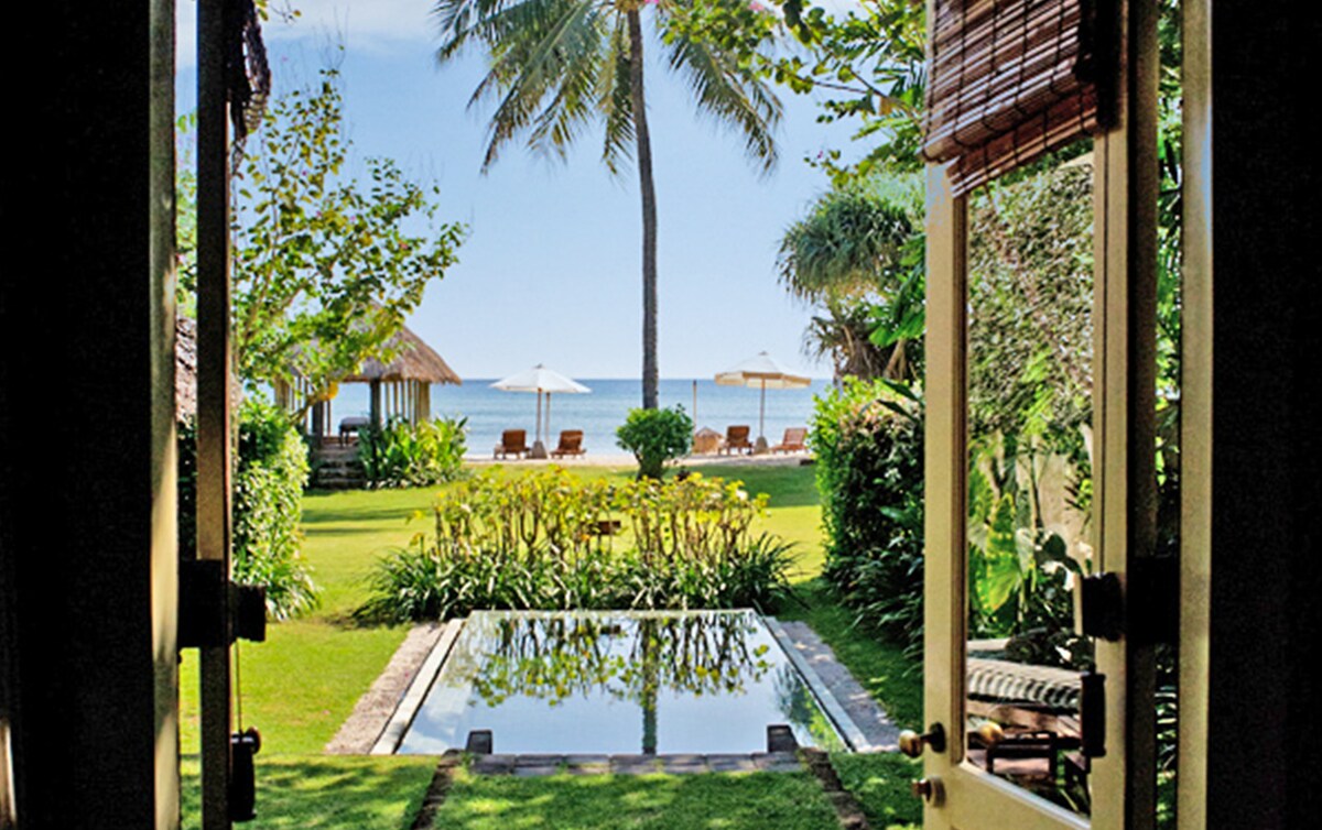 Double doors open to reveal a private tropical garden featuring lush greenery and a small plunge pool. In the background, the ocean is visible, framed by palm trees and sun loungers under umbrellas, creating a serene outdoor space.