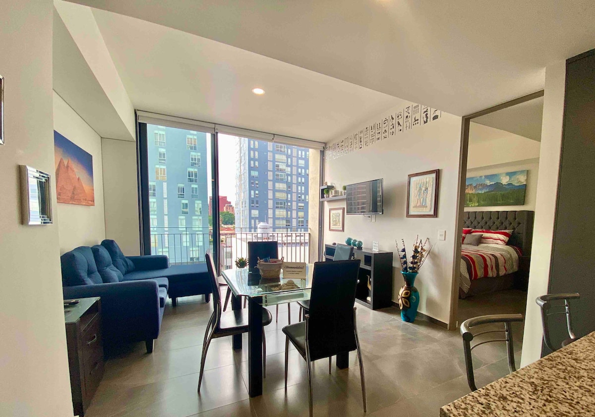 An open-concept living area showcases a modern dining table surrounded by six chairs. A blue sofa and a media console are positioned nearby, with large windows offering ample natural light and views of the surrounding buildings.