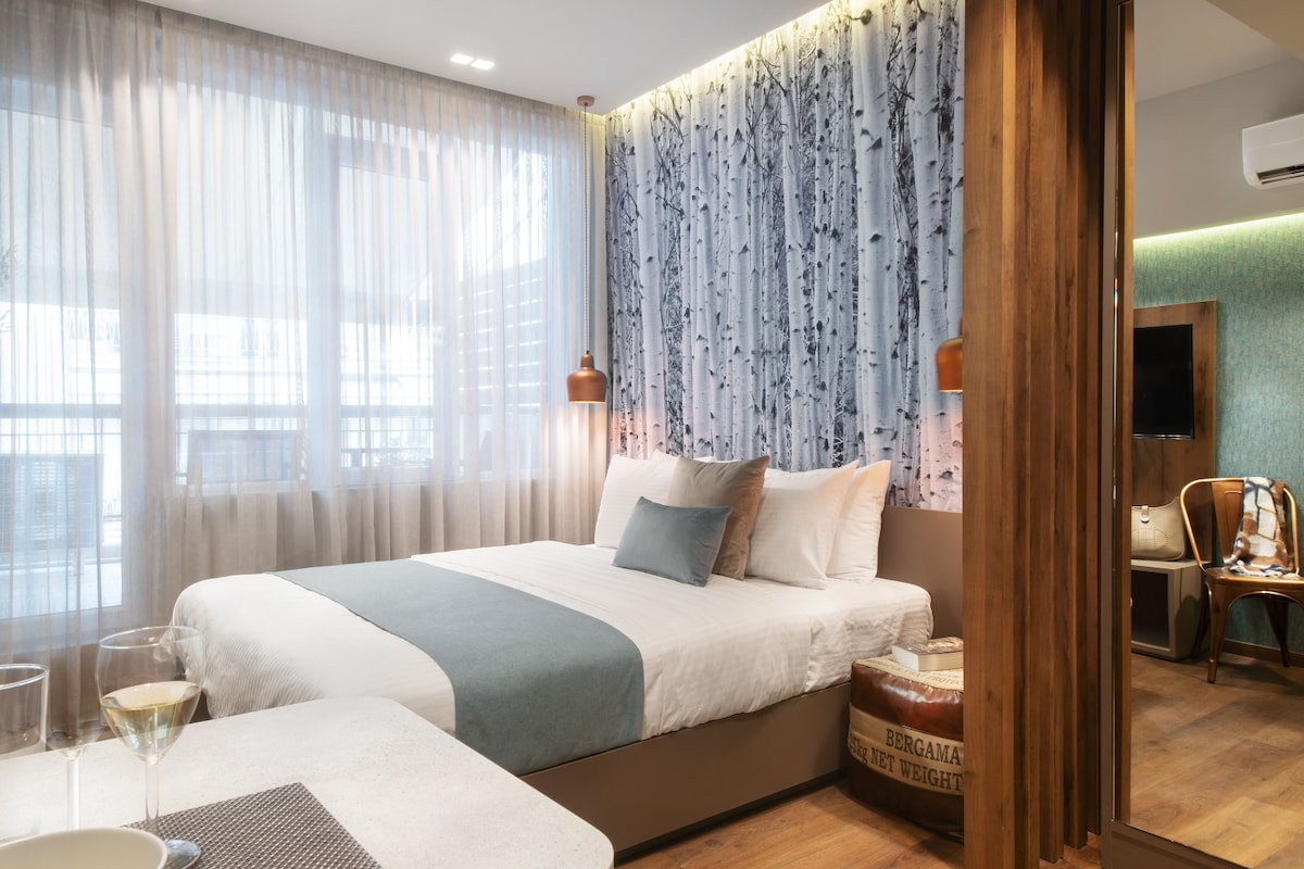 A modern studio is highlighted by a large window draped with sheer curtains, allowing natural light to filter softly in. A spacious bed features a light blue accent blanket, complemented by neutral-colored pillows. Wooden elements add warmth to the contemporary decor, while a muted wall art design evokes a serene outdoor ambiance.