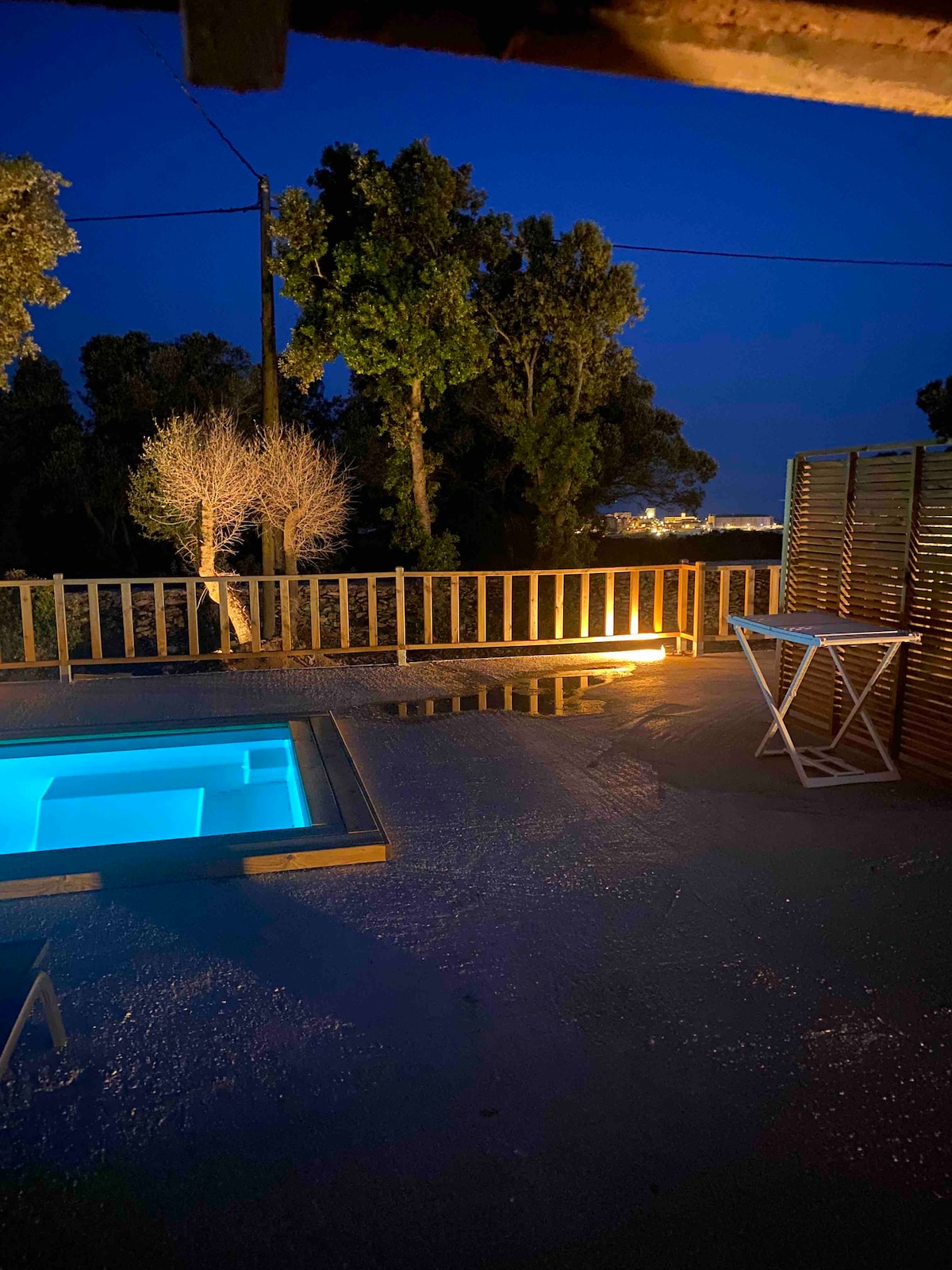 The evening scene features a private heated pool illuminated in soft blue light, surrounded by wooden decking. A small table and chairs are positioned nearby, with trees framing the view in the background, hinting at the peaceful ambiance of the location.
