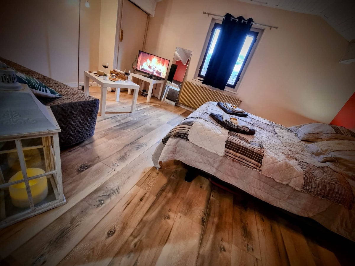 A cozy studio features a spacious bed covered with a textured blanket. A neutral palette is complemented by wooden flooring. A seating area is arranged with a low table, and a television is visible displaying a fireplace scene. Large windows allow natural light, with dark curtains partially drawn.