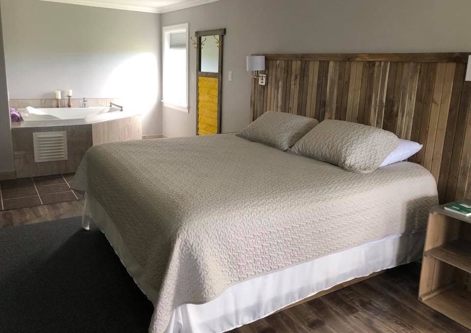 A spacious room features a king-sized bed topped with a light quilt and two pillows. A jacuzzi tub is positioned nearby, while natural light filters through a window, adding warmth to the earthy-toned decor. A wooden headboard and bedside lamps enhance the inviting ambiance.