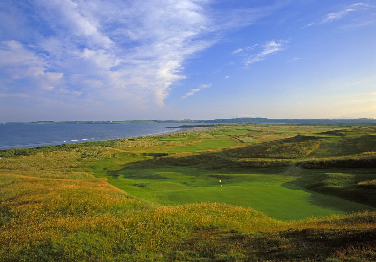 An expansive view of a coastal golf course is displayed, featuring lush green fairways that lead toward the shimmering blue waters of the ocean. The surrounding landscape includes gently rolling hills and a clear sky, symbolizing a serene and inviting outdoor environment.