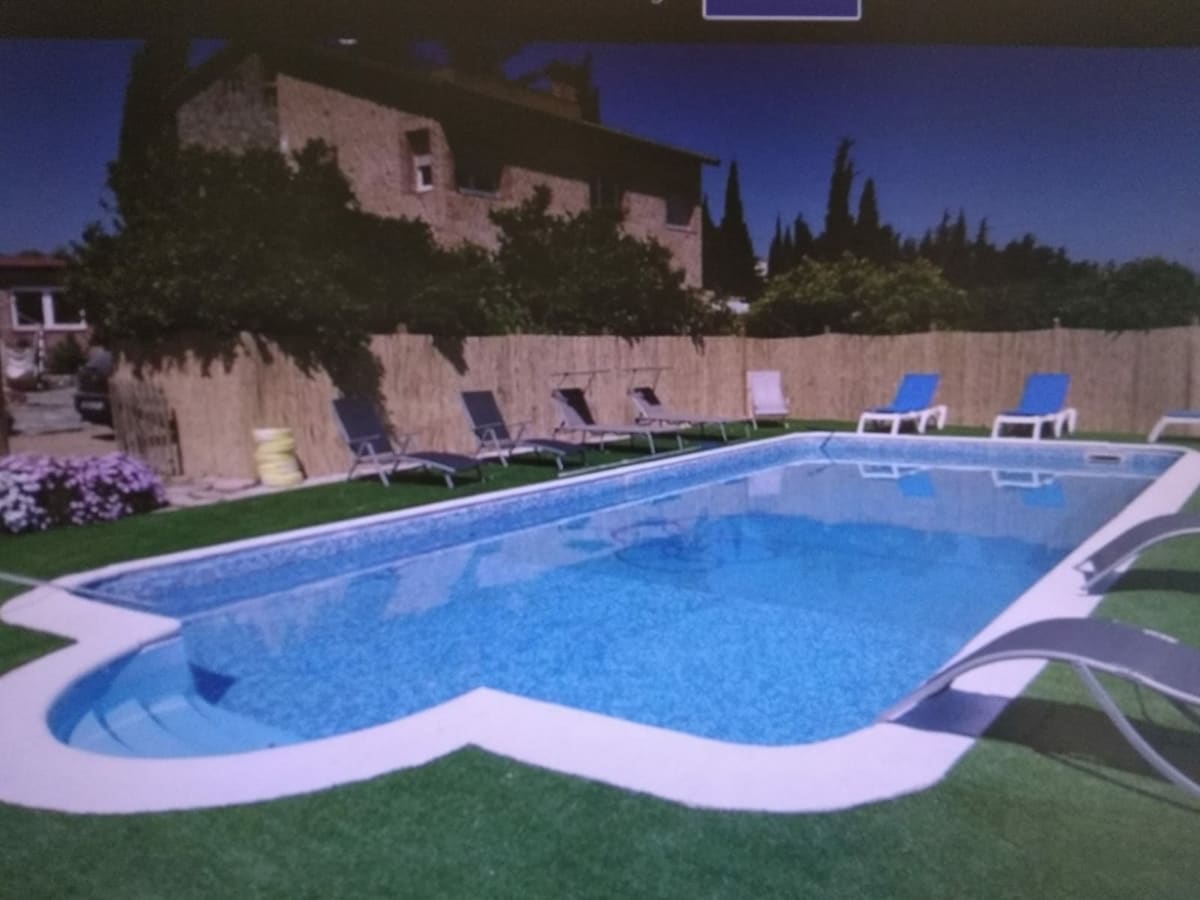 A large, well-maintained swimming pool is surrounded by a grassy area and enclosed by wooden fencing. Several sun loungers in varied colors are positioned around the pool, providing ample space for relaxation. Lush green plants and flowers add to the outdoor ambiance.