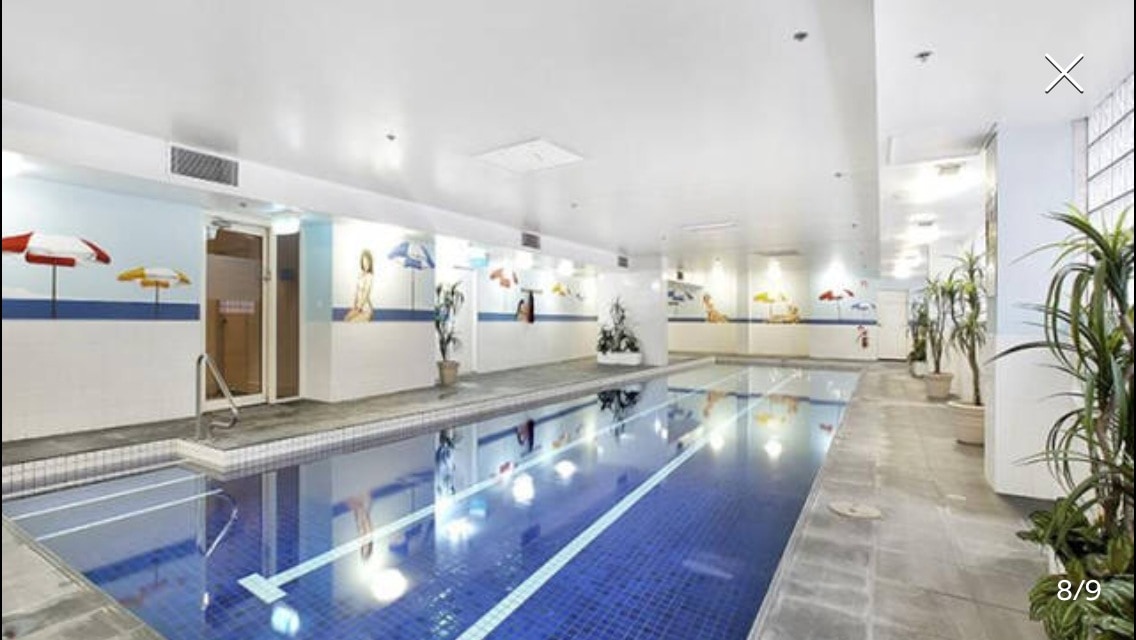An indoor swimming pool is surrounded by bright artwork depicting colorful beach umbrellas. Natural light is provided by overhead fixtures, reflecting off the tranquil water. Potted plants add a touch of greenery to the spacious area, enhancing the leisure atmosphere.