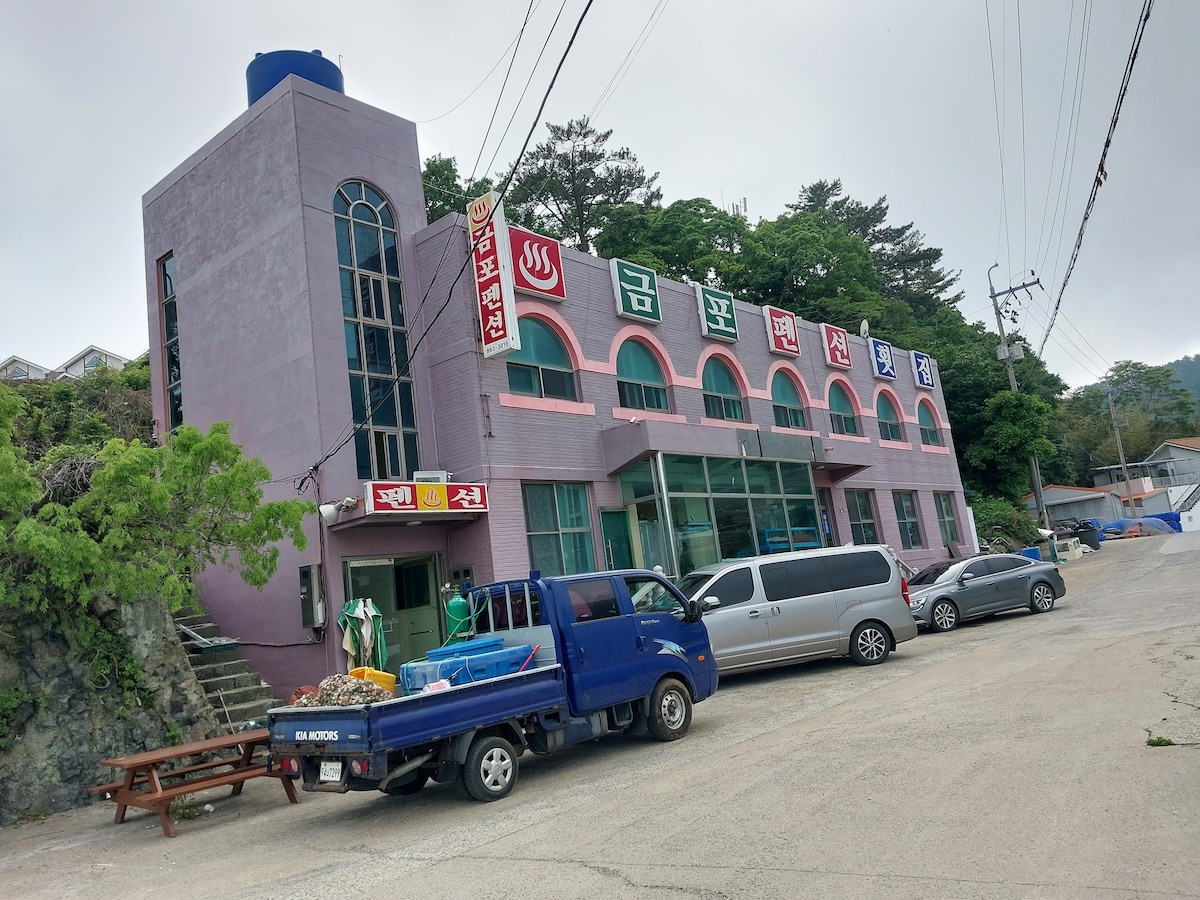 A multi-story building is showcased with a pink exterior and large, arched windows. The facade features signs displaying the establishment's name. A parking area is visible in front with several vehicles, including a blue truck and a gray minivan. Surrounding greenery adds a natural touch.