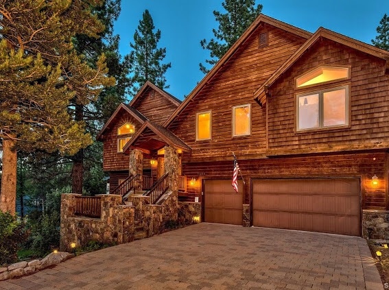 The exterior of the custom-built Alpine lodge features rustic wooden siding and rugged stone accents. Warm lights glow from the windows, enhancing the inviting appearance. A spacious driveway leads to a three-car garage, surrounded by tall trees and a serene atmosphere.