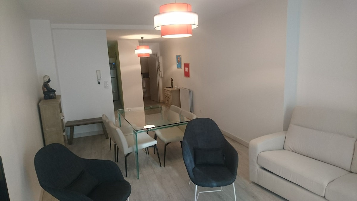 A spacious living area is presented with a glass dining table surrounded by six chairs. Two accent chairs are positioned nearby, and a light-colored sofa is situated against the wall. Soft pendant lighting hangs from the ceiling, enhancing the bright and airy ambiance.