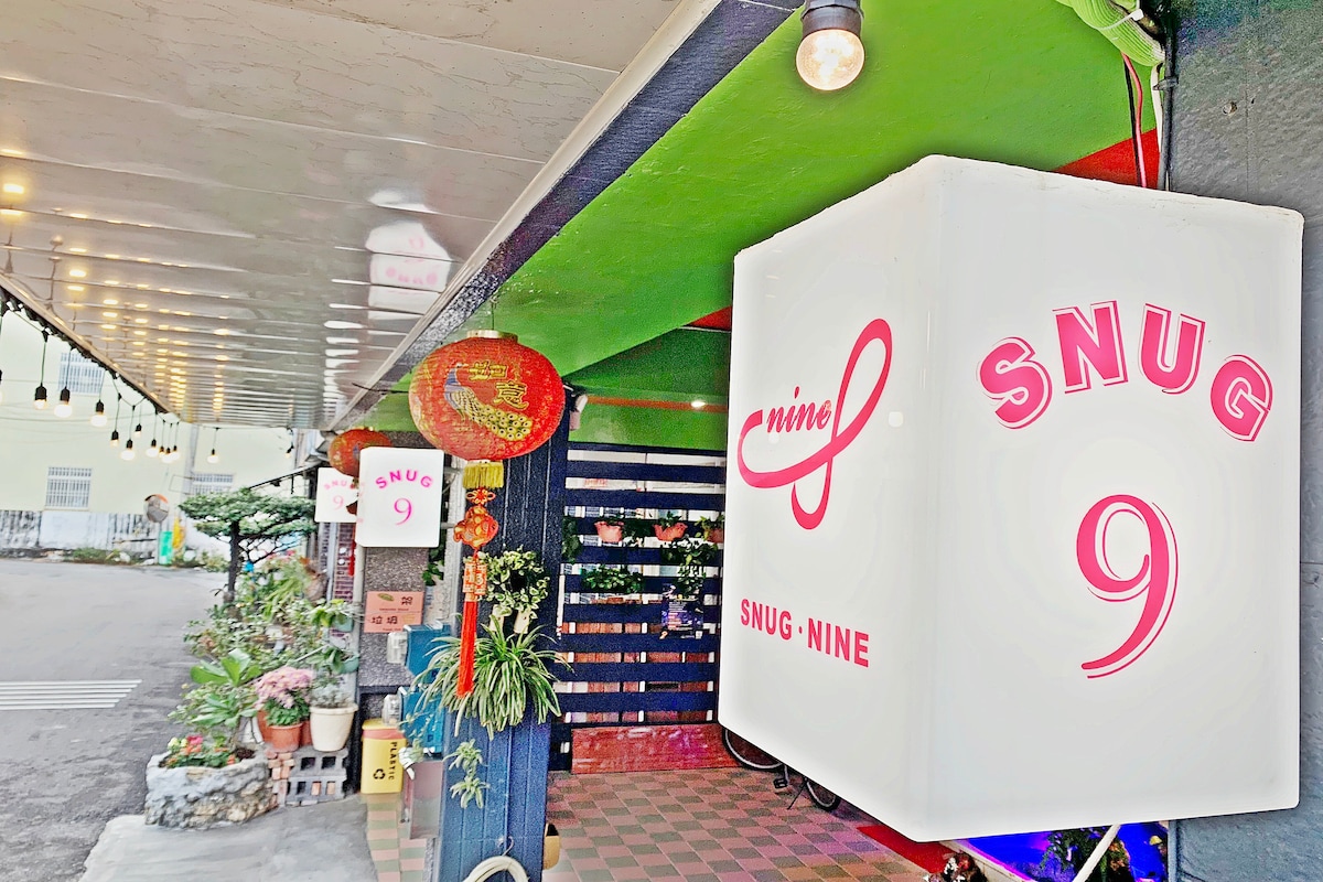 The entrance features a colorful façade with the 'SNUG 9' sign prominently displayed. Potted plants add a touch of greenery along the walkway, and decorative paper lanterns hang from the ceiling, creating a welcoming vibe.