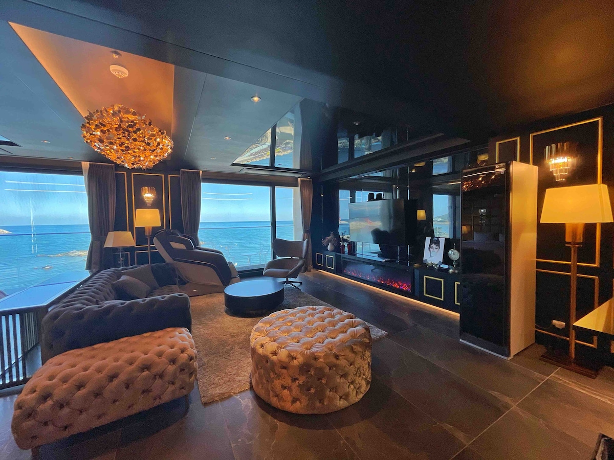 A modern living area features large windows offering ocean views and natural light. Luxurious furnishings include a plush sofa, a stylish round ottoman, and a sleek media console. Light fixtures add a sophisticated touch against the dark wall accents.