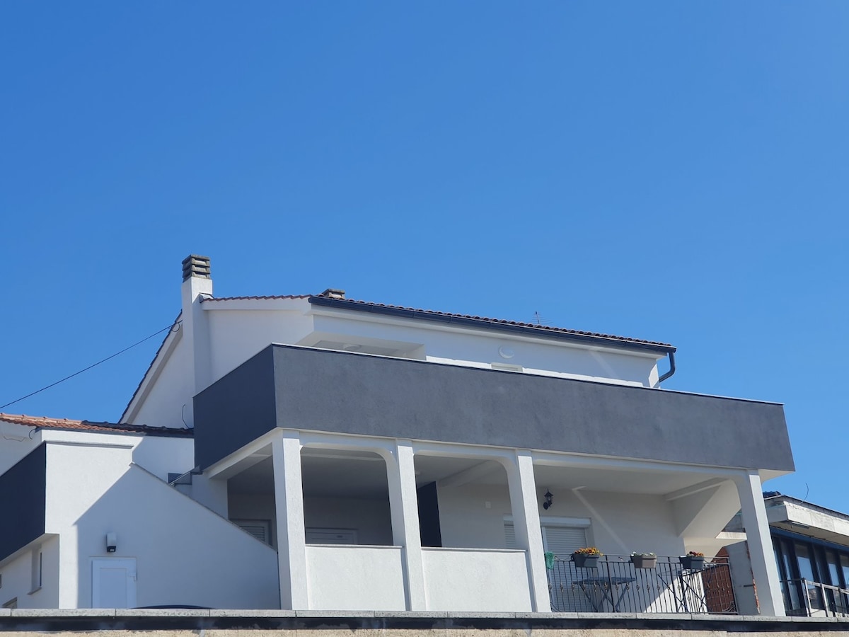 A modern two-story apartment building is set against a clear blue sky. The structure features a large balcony with a railing, enhancing the overall aesthetic. Clean lines and neutral colors characterize the architecture, creating a contemporary appearance.