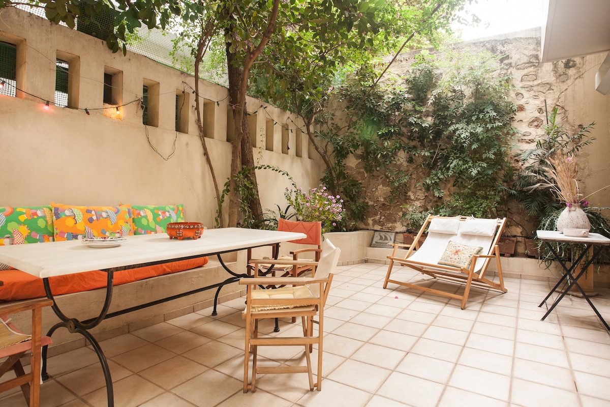 A spacious private courtyard is surrounded by greenery, featuring a large rectangular table with chairs, and a reclining lounge chair. Soft lighting accents the area, while vibrant cushions line one side of the table, creating a relaxing environment for outdoor gatherings.