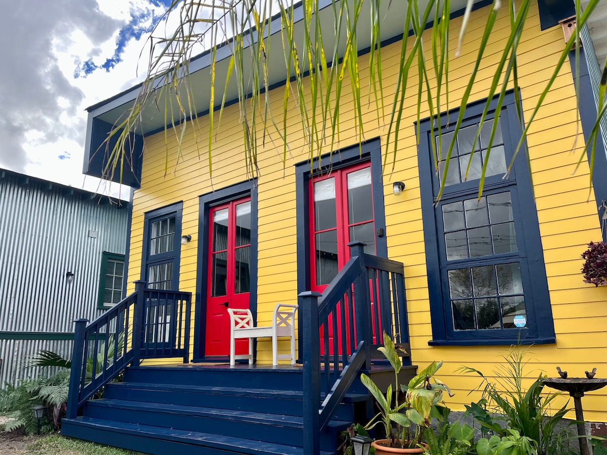 A charming two-story exterior showcases vibrant yellow siding with bold red doors and blue trim. The entrance is framed by a welcoming staircase and lush tropical plants, inviting guests into a warm space filled with character.