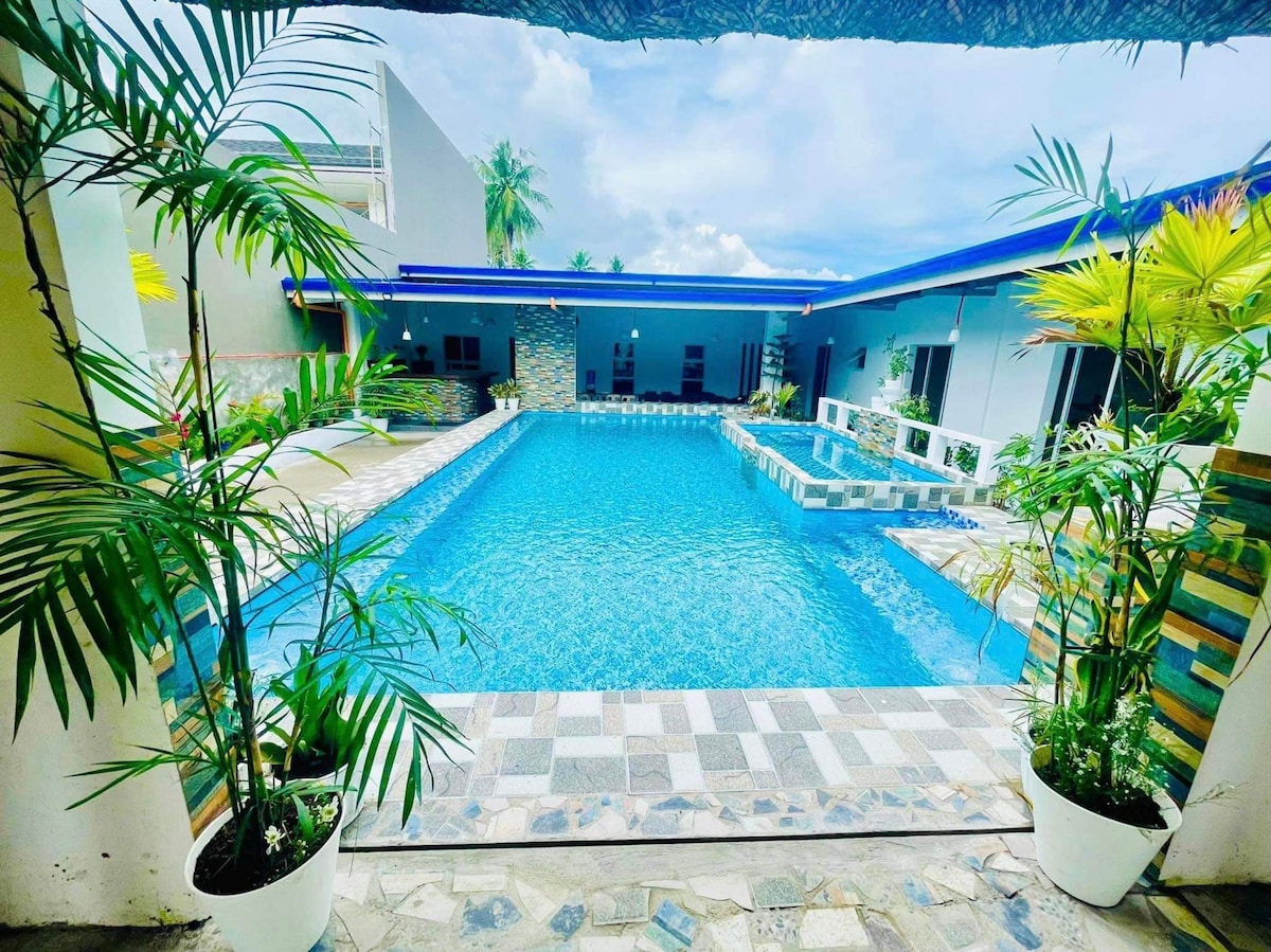A private hot spring pool is surrounded by a stone-paved area, featuring several lounge spaces. Lush green plants in white pots frame the pool, while a modern villa is visible in the background, showcasing open spaces and a clear blue sky.