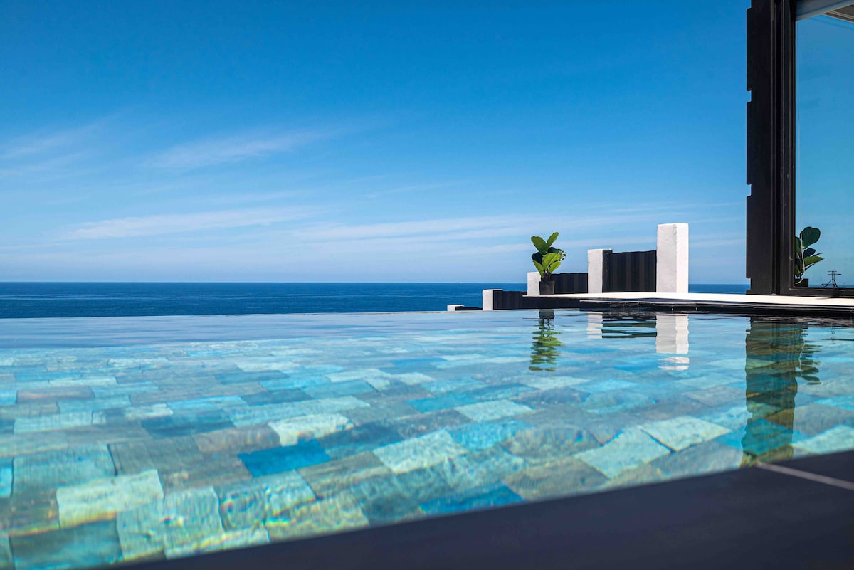 A serene glass pool extends towards the horizon, featuring turquoise tiles that reflect the sky above. The ocean can be seen in the background, with gentle waves meeting the shore. Minimalist planters flank the edge, enhancing the peaceful ambiance of the space.