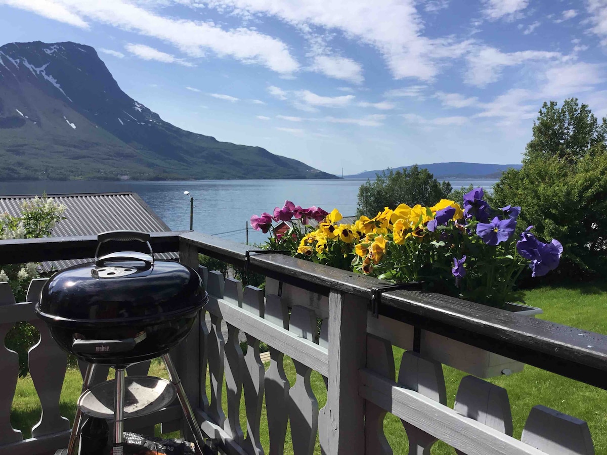 A balcony view features vibrant flower pots filled with yellow, purple, and blue pansies. A small grill is positioned nearby, with panoramic views of the sea and distant mountains under a clear blue sky, enhancing the outdoor ambiance.
