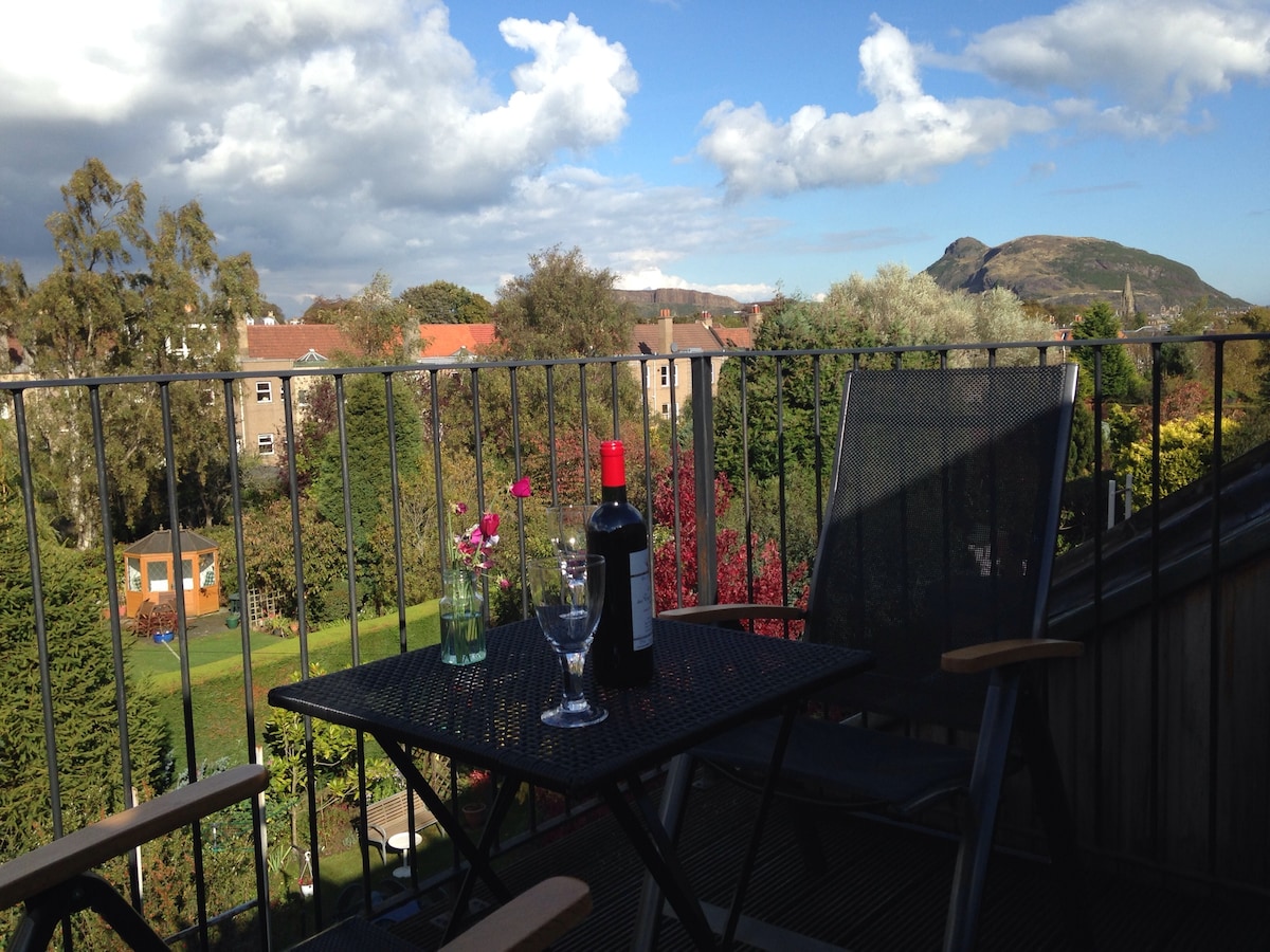 A small table with a bottle of red wine and a glass is set on a private terrace, surrounded by greenery. A view of a distant hill and clouds is visible, enhancing the serene outdoor space. Two chairs provide seating for relaxation.
