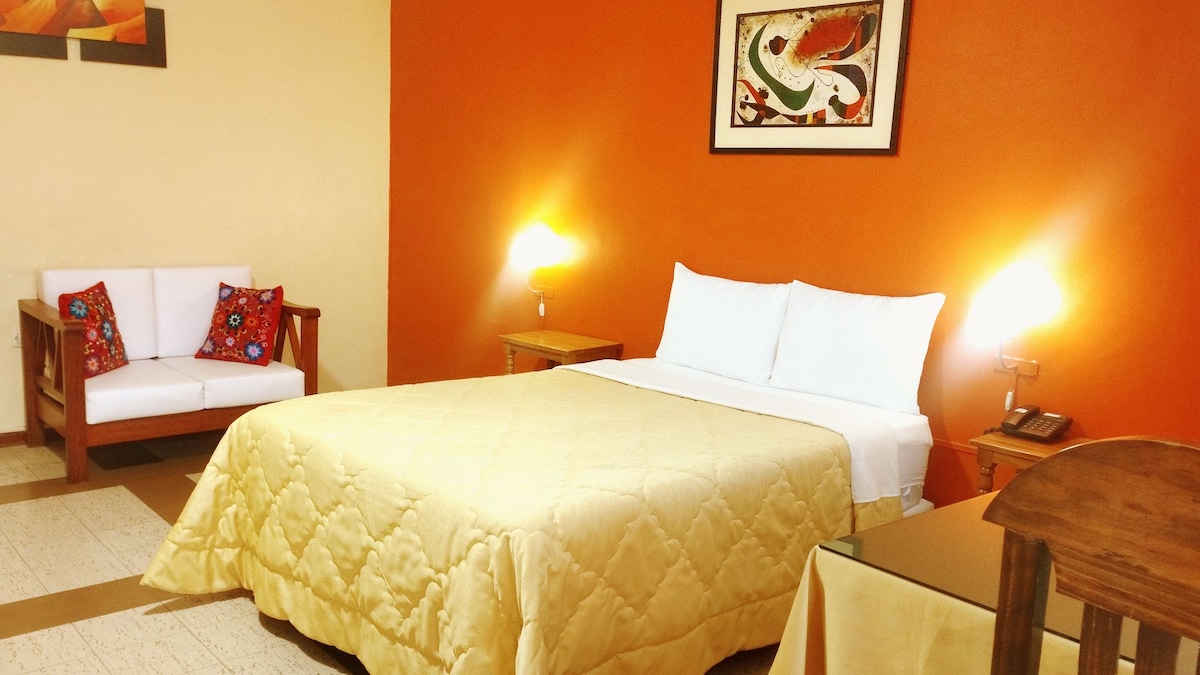 A cozy room features an inviting yellow bedspread on a spacious bed. An orange accent wall adds warmth. A small seating area with decorative pillows is visible, along with a wooden table and a light fixture, providing adequate illumination.