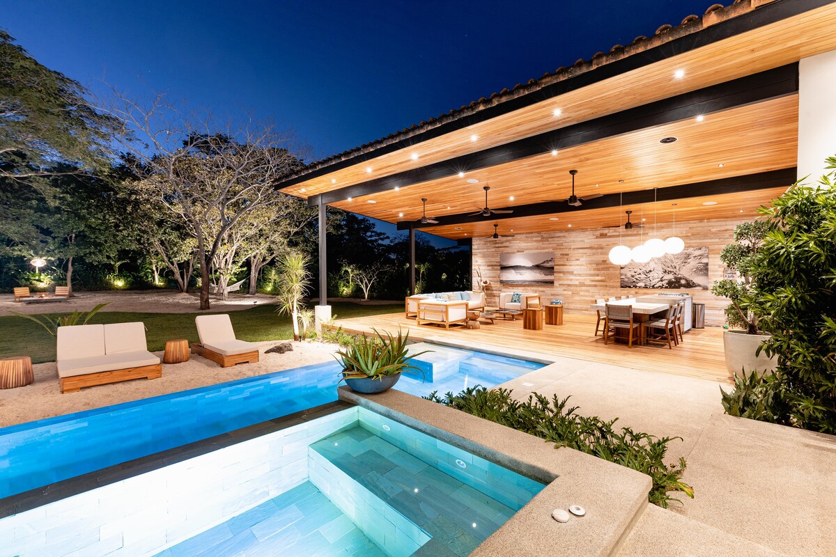 An outdoor area featuring a non-heating jacuzzi and a small pool, illuminated in soft blue lighting. A spacious covered patio provides comfortable seating and a dining area, surrounded by lush greenery and trees. The space combines modern architecture with natural elements.