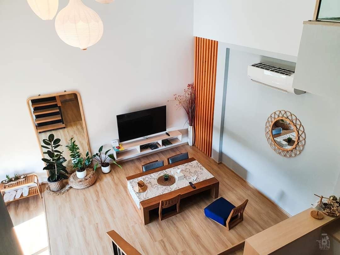 An airy living area features a large mirror, potted plants, and warm wood accents. A marble-topped table is surrounded by wooden chairs, while a flat-screen TV is mounted on the wall. Natural light filters in through high windows, complementing the open design.