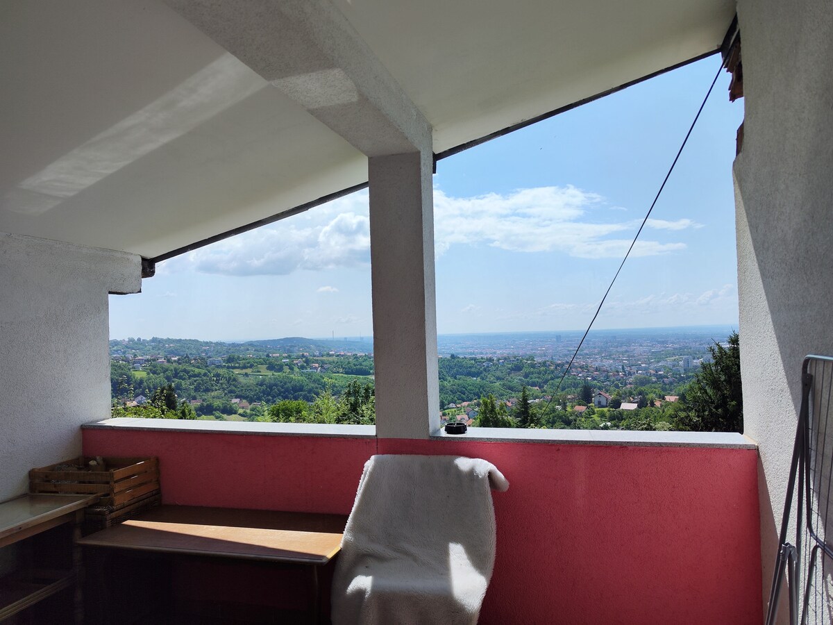 A spacious balcony is framed by two walls, offering panoramic views of the surrounding landscape. Natural light floods the area, highlighting a wooden table beside a cozy chair. The scene showcases a serene environment with lush greenery and distant city views under a clear blue sky.