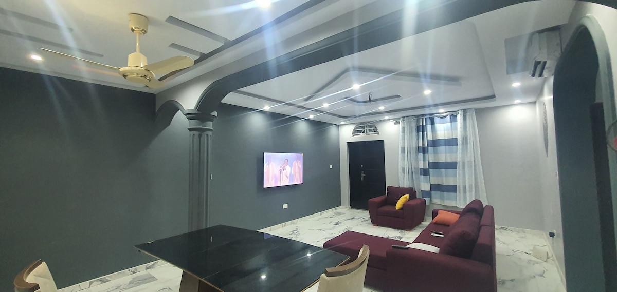 A spacious living area features a dark grey accent wall, complemented by a sleek black dining table and plush maroon sofas. A large TV is mounted on the wall, providing a focal point. Bright ceiling lights and a ceiling fan enhance the modern interior.