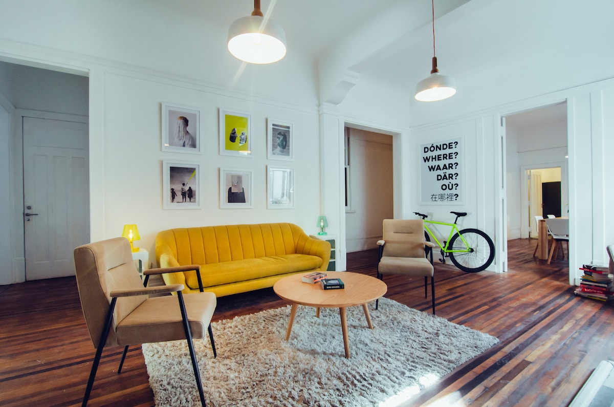 A spacious living area features a vibrant yellow sofa complemented by two neutral armchairs. A round coffee table sits on a textured rug, and a bicycle leans against the wall. Decorative artwork frames the walls, enhancing the modern aesthetic of the room.