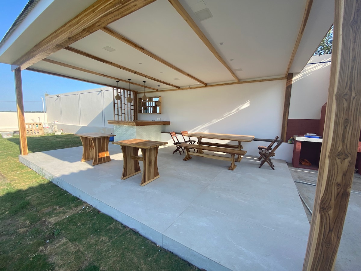 An outdoor area is showcased featuring a covered space with wooden furniture, including a large table and benches. The floor is finished with a smooth surface, and light streams in, creating a bright and open atmosphere. A rustic bar is visible in the background.
