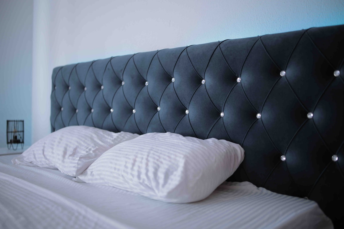 A cozy sleeping area features a tufted headboard with decorative buttons, complementing the crisp white linens on the spacious bed. Natural light fills the space, enhancing the serene ambiance of the room.