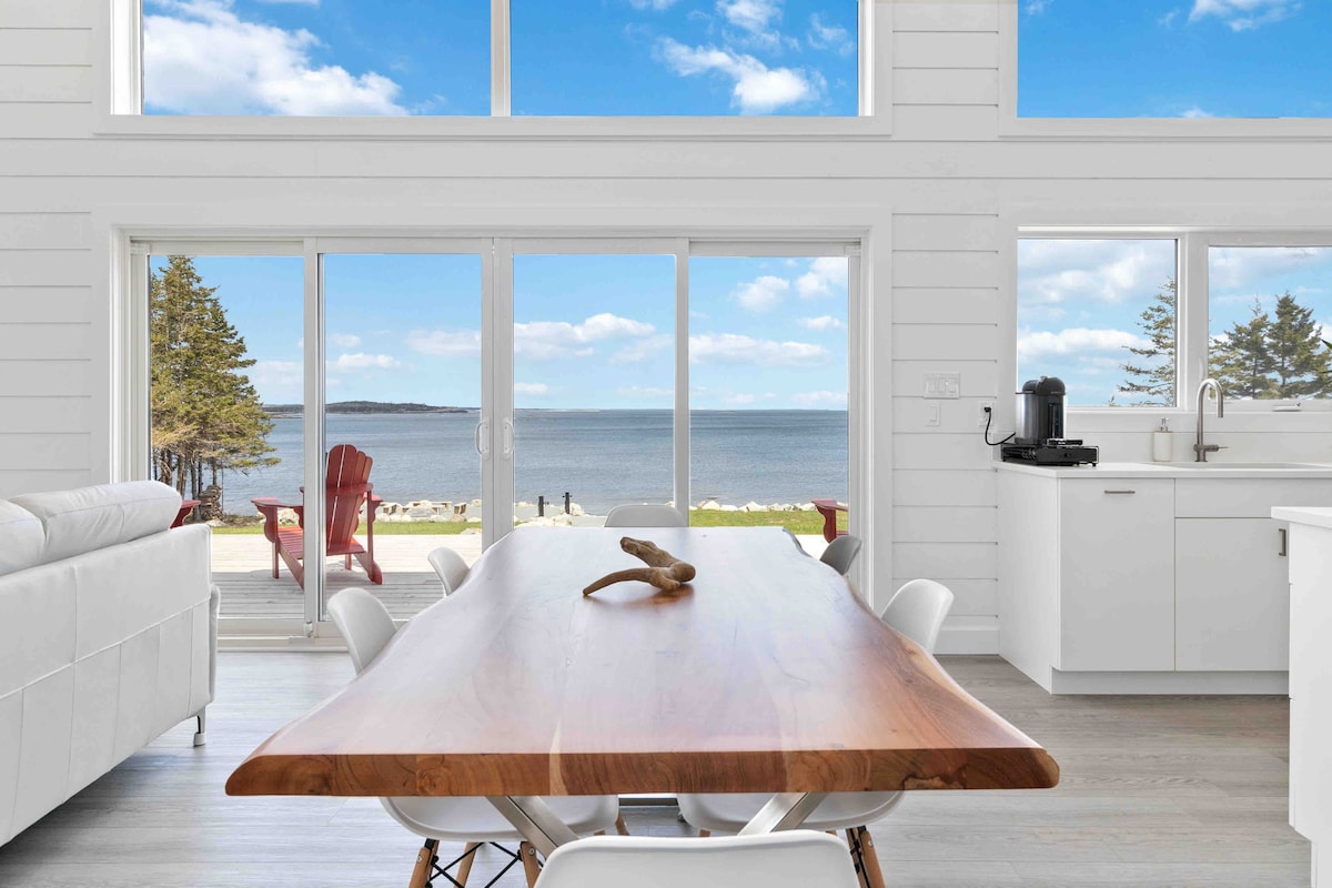 A spacious dining area features a long wooden table surrounded by white chairs, with large windows offering panoramic ocean views. Natural light fills the room, enhancing the clean lines and modern design. The serene outdoor landscape is visible through sliding glass doors.