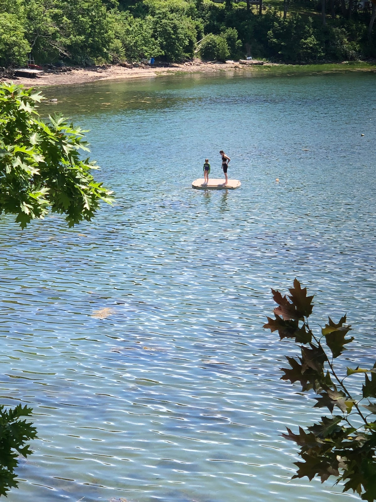 Two individuals are seen standing on a floating platform in a calm, shimmering body of water. Green foliage frames the image, highlighting the serene natural setting. The sun casts gentle reflections on the water's surface, suggesting a warm, inviting atmosphere.