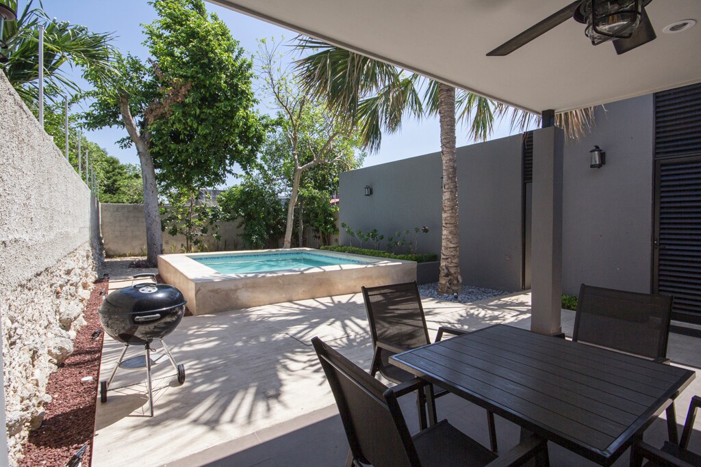 A sunlit patio area showcases a private pool surrounded by lush greenery, featuring palm trees and tropical plants. A charcoal grill is positioned near a dining table with seating for four, providing an inviting space for outdoor gatherings.