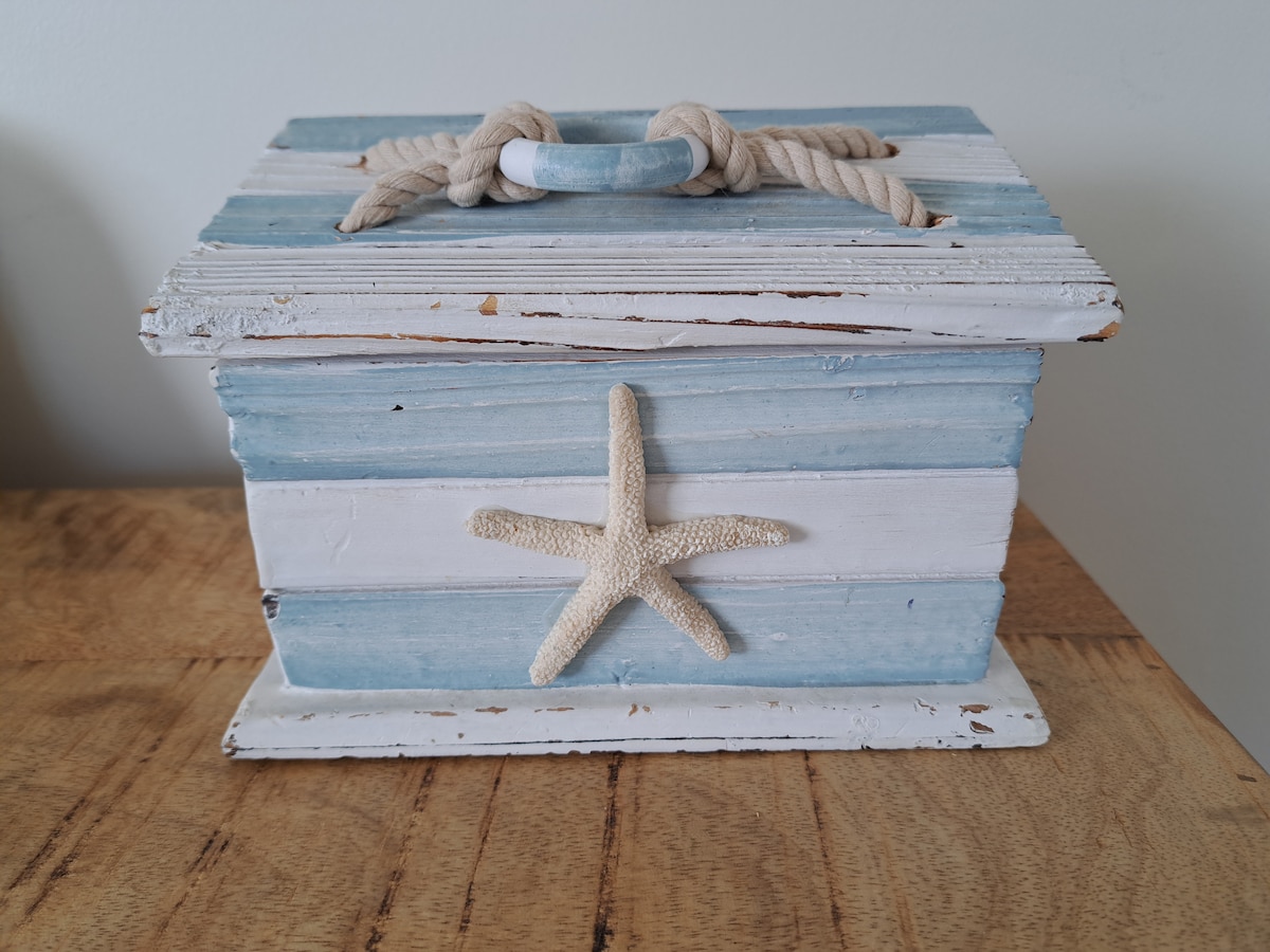 A wooden storage box is displayed with a weathered finish in soft blue and white stripes. A starfish decoration is affixed to the front, and a rope handle is attached to the lid, enhancing its coastal theme.