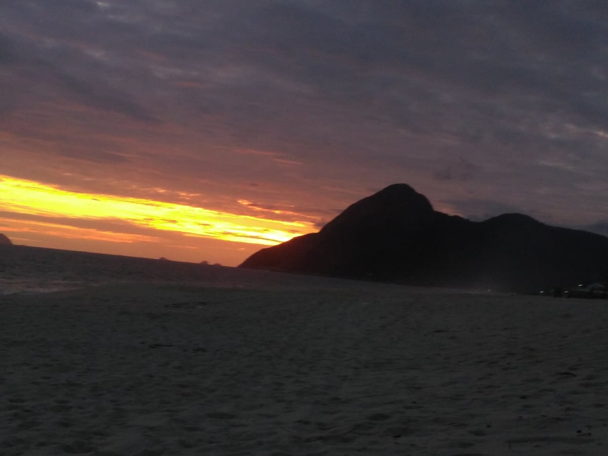 A beach scene captured during sunset, showcasing a gradient of warm colors in the sky. The silhouette of a dark mountain rises in the background, while gentle waves lap against the sandy shore. The horizon is marked by distant outlines of land.