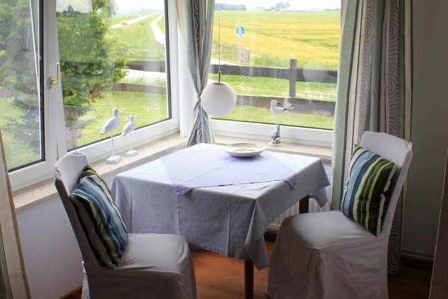 A bright dining area features a small white table draped with a light purple cloth. Two chairs with striped cushions are positioned around the table. Large windows offer scenic views of the surrounding fields and greenery, allowing natural light to fill the space.