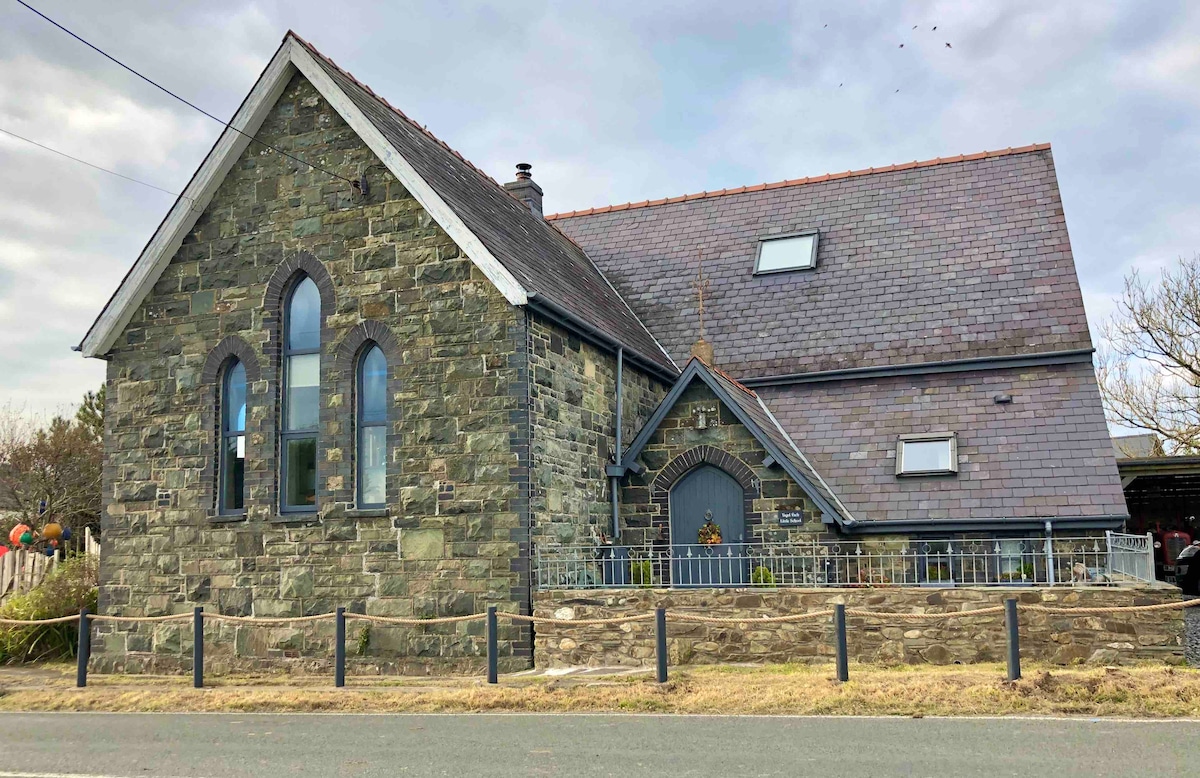 The exterior of a historic stone building is showcased, featuring large gothic style windows and a colorful garden. A sloping roof with multiple skylights is visible. The property is surrounded by a low stone wall and a gravel pathway leads to the entrance.