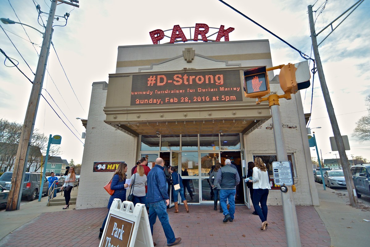 The image shows the entrance of the Park Theater, a bright marquee displaying 'D-Strong' invites attendees for a fundraising event. A crowd is seen entering the building, with a mix of individuals casually dressed. Streetlights and utility poles frame the scene, contributing to the urban setting.