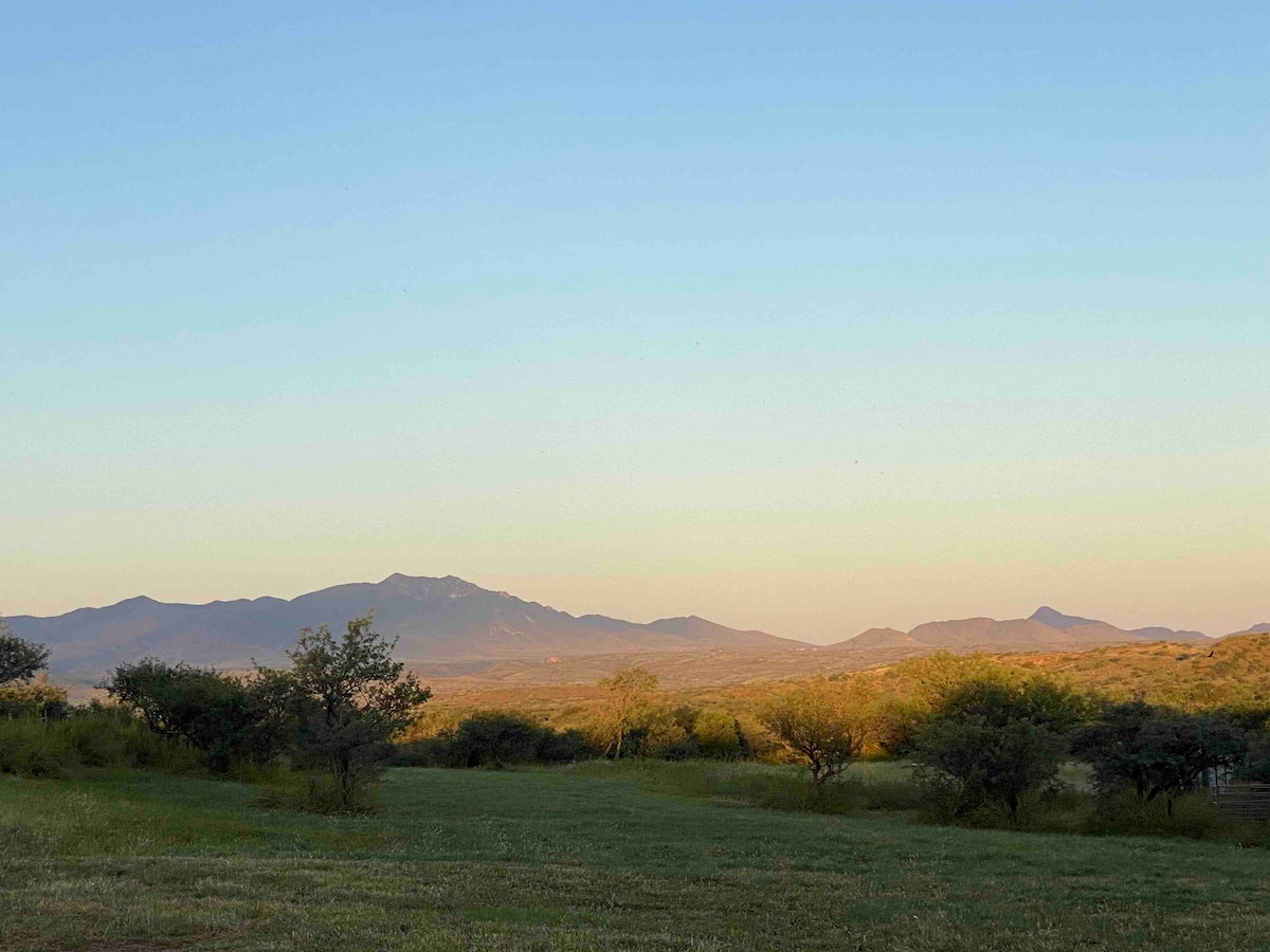 A panoramic view of rolling hills and mountains is visible under a clear sky at dusk. The landscape features gentle slopes with patches of grass and scattered trees, offering a serene and natural setting.