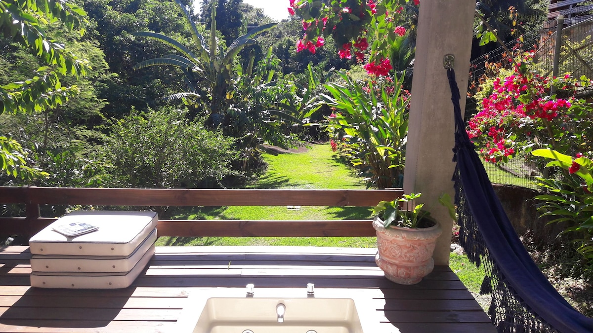 A private deck overlooks a lush garden filled with vibrant flowers and greenery. A hammock hangs nearby, inviting relaxation. A non-heated hot tub is positioned on the deck, providing a serene spot for enjoyment under the sun.