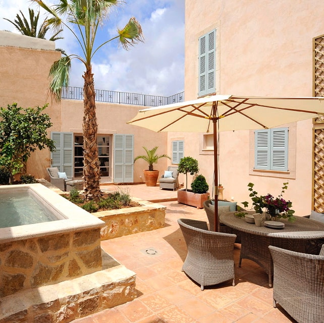 A tranquil patio area is presented, featuring stone walls that complement the terracotta flooring. A shaded dining table with woven chairs sits next to a inviting pool, surrounded by lush greenery and potted plants. Sunlight filters through the surroundings, creating a serene atmosphere.