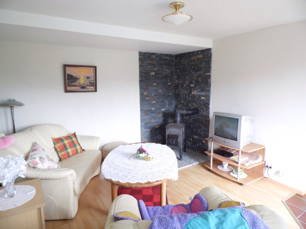 A comfortable living area features a light-colored sofa and a cozy armchair. A round table adorned with a lace tablecloth is positioned centrally. A small stove is located against a stone accent wall, complementing the soft natural light from the room.