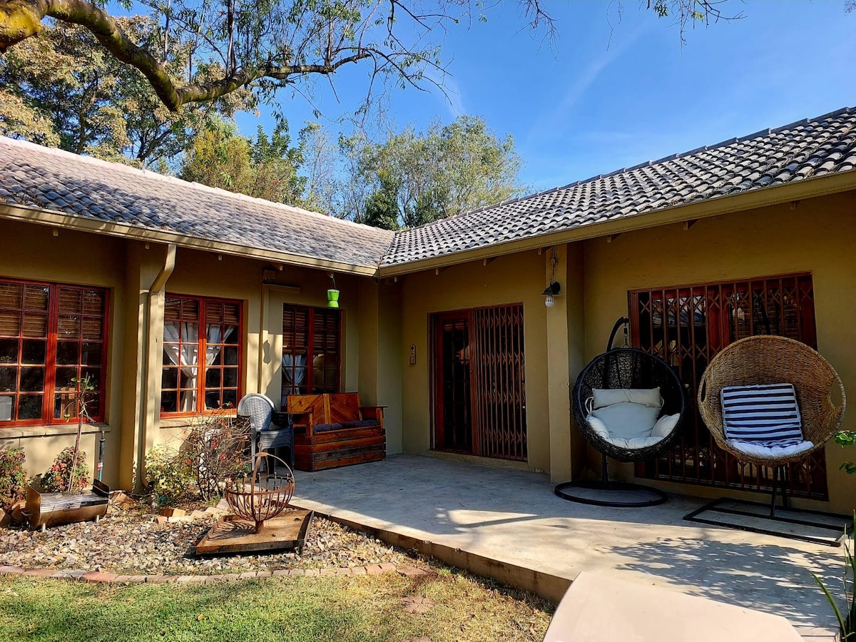 A spacious patio area features two hanging chairs and a wooden bench, surrounded by neatly arranged gravel and plants. The light-colored building is complemented by wooden window frames and a gently sloping roof, with abundant greenery visible in the background.