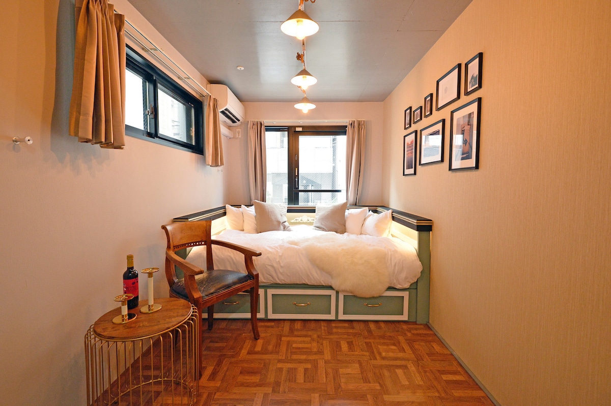A cozy sleeping area features a spacious bed adorned with white bedding and plush pillows. Natural light filters through large windows, complemented by curtains. A small wooden side table holds a bottle, and framed photographs line the wall, adding character to the room.